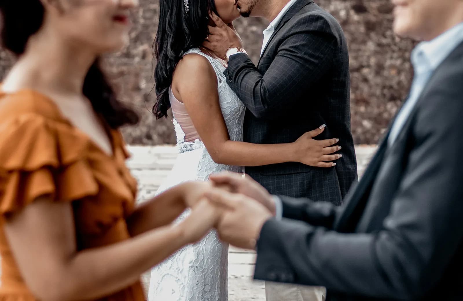 Noivos se beijam enquanto dois convidados dão as mãos em primeiro plano