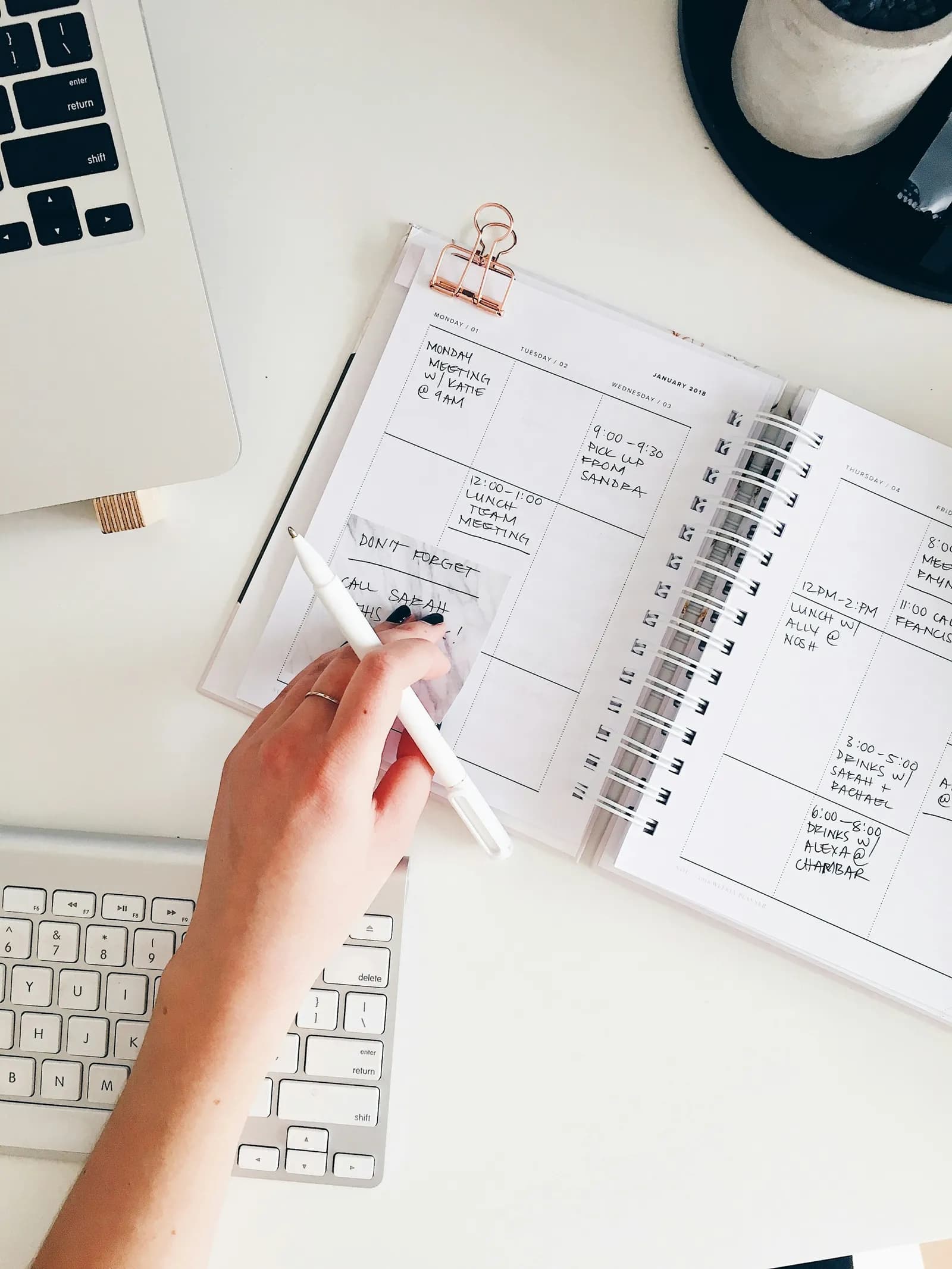 Mão anotando em uma agenda aberta sobre a mesa de trabalho